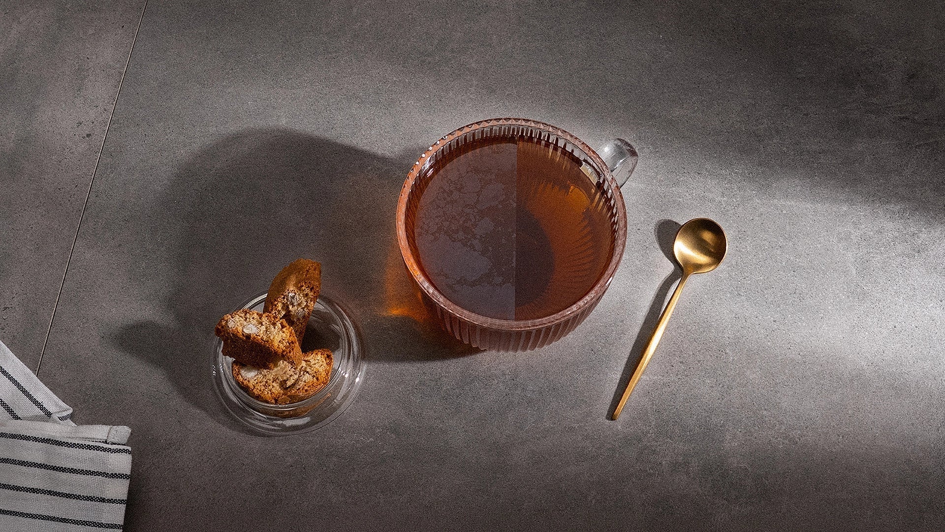 A cup of tea with two sides: one with a film on top and one without. Next to is lies a spoon and on the other side stands a bowl with cookies.