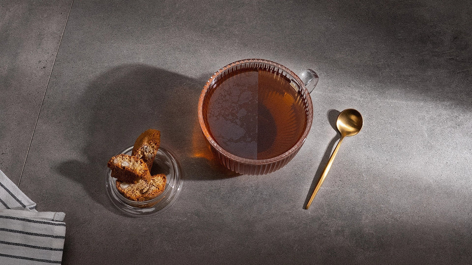 A cup of tea with two sides: one with a film on top and one without. Next to is lies a spoon and on the other side stands a bowl with cookies.