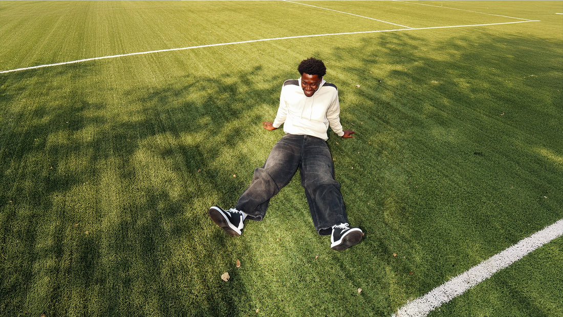 A young man sitting on a grass sports field, wearing a light-coloured hoodie, dark jeans and black trainers, with white field markings visible in the background.