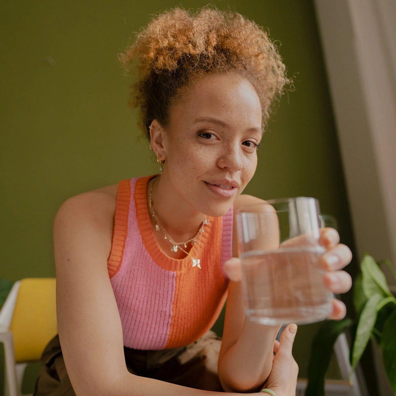 Woman in a pink top holding a glass of water in a home setting with plants.