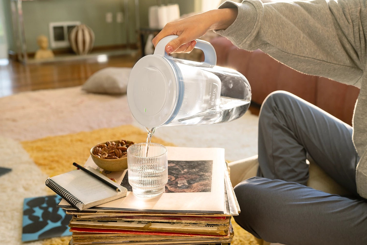 Person pouring filtered water from a blue BRITA Glass Jug into a drinking glass in a living room.