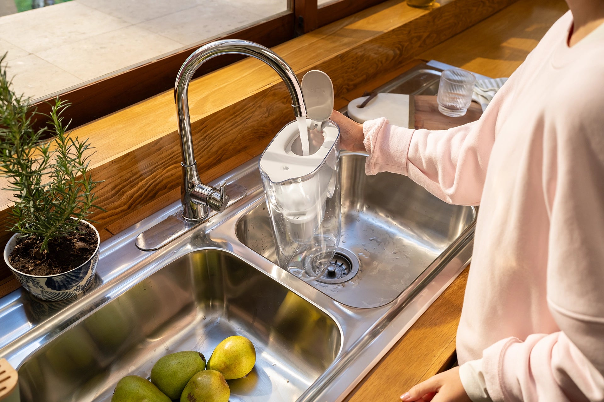 Person filling a white BRITA Style Essential with tap water at a kitchen sink, with fresh fruit in the sink.