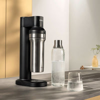 Black BRITA sodaTRIO Sparkling Water Maker with a BRITA sodaTRIO Glass Bottle and a glass of sparkling water on a countertop.