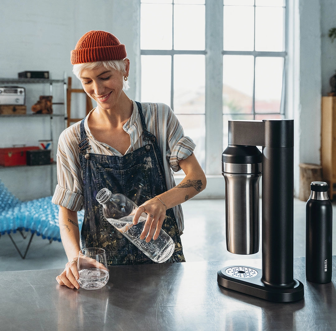 Woman pouring sparkling water into a glass beside a BRITA sodaTRIO sparkling water maker on a kitchen counter.