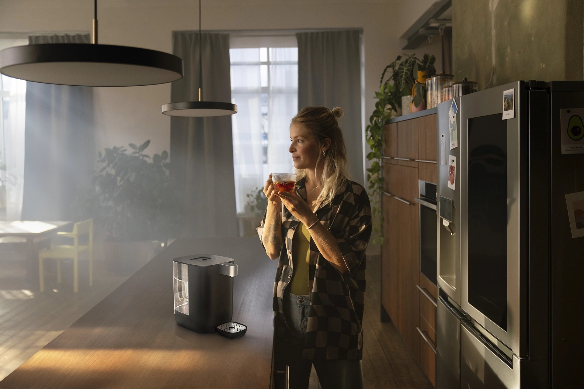 Woman holding a glass cup of tea in a kitchen next to a BRITA YOURCE mini water dispenser on the counter.